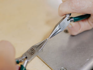 Close-up of a hand using 2 pairs of chain nose pliers on a metal surface | jewelry making pliers