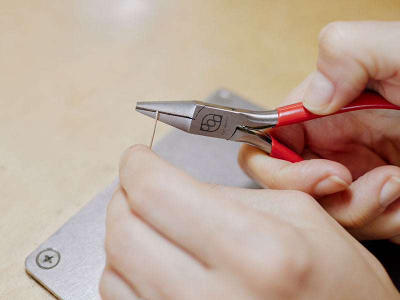 Close-up of hands using a pair of chain-nose pliers on a light-colored surface | jewelry pliers