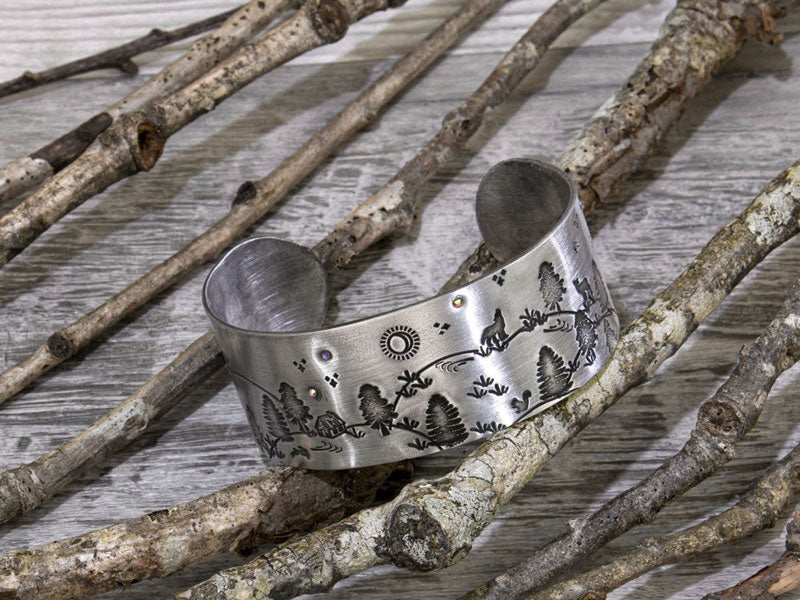 A silver metal bracelet with embossed patterns of trees and nature, placed on a background of wooden branches.